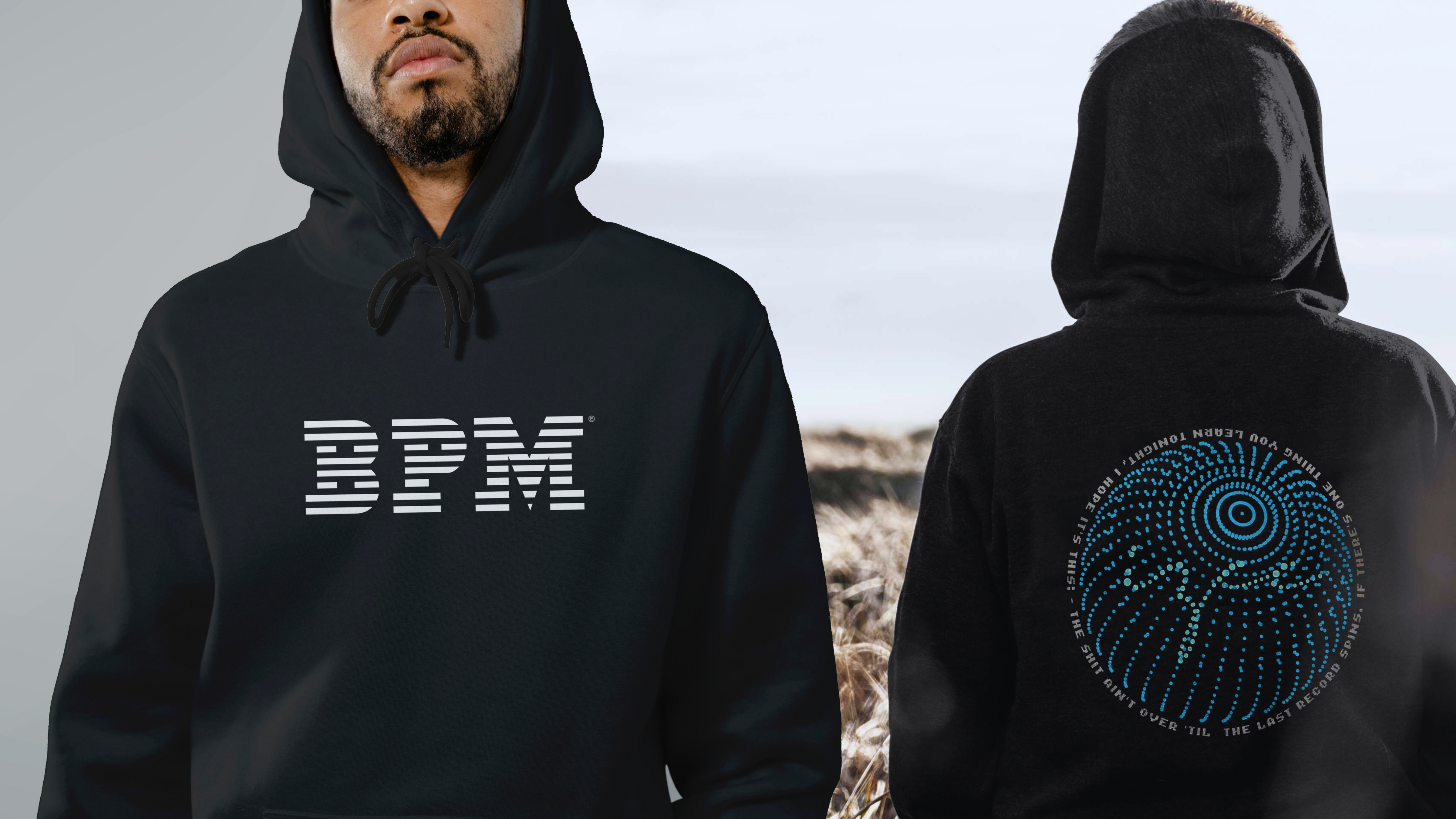 A stylish promotional image featuring two individuals wearing black hoodies with unique designs inspired by music culture. The person on the left is shown from the front, wearing a black hoodie with a bold, modern white "BPM" logo across the chest, where each letter is formed using horizontal stripes, evoking a sense of rhythm and movement. The person on the right is shown from behind, standing outdoors in a field of golden wheat under a cloudy sky. Their hoodie features a vibrant, circular blue graphic resembling a vinyl record or sound wave pattern, made of concentric rings and dots. Encircling the graphic is a quote in white uppercase text that reads: “IF THERE’S ONE THING YOU LEARN TONIGHT, I HOPE IT’S THIS: THE SHIT AIN’T OVER ‘TIL THE LAST RECORD SPINS.” The overall aesthetic of the image conveys a cool, modern vibe with strong connections to electronic music and DJ culture, blending urban fashion with a love for sound and rhythm.